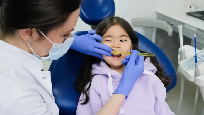 kontrola-zebow-mlecznych-u-dziecka Dentystka w rękawiczkach bada zęby uśmiechniętej dziewczynki w gabinecie.